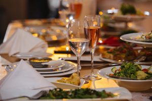 Relatively long table at low angle, dishes and wineglasses served for an upcoming banquet event. Salad, soup, casserole, variety of wine. Triangularly folded napkins. Blur behind focal point.